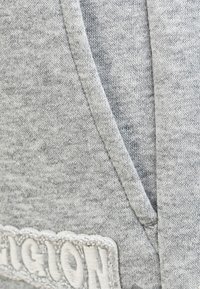 Jasnoszary materiał dresowy o miękkiej teksturze, z dekoltem w kształcie litery V i białym haftowanym logo. Widoczny delikatny melanż.