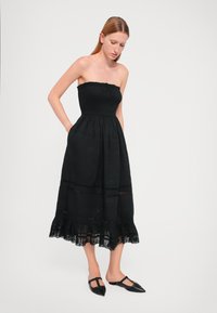 Vestido maxi preto sem alças com tecido texturizado, corpete franzido, bolsos laterais e bainha com rendas. Combinado com sapatilhas pretas de bico fino.