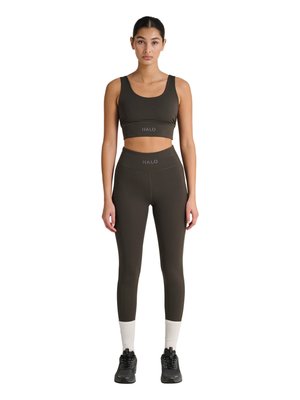 Vrouw staat en draagt bijpassende donker olijfgroene sportbeha en leggings met hoge taille met "HALO"-logo, witte sokken en zwarte sportschoenen.