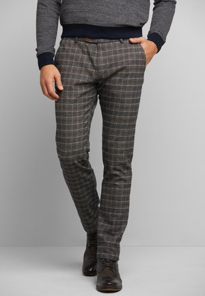 Mann trägt graue karierte Hose, dunkle geschnürte Lederschuhe und einen grauen Pullover mit dunklen Bündchen, steht vor einem schlichten Hintergrund.