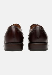 Scarpe eleganti in pelle marrone con dettagli brogue, punta tonda e tacco in legno impilato. Texture liscia con intricate perforazioni lungo le cuciture.