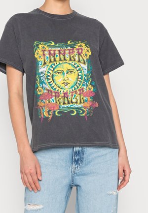Osoba nosząca wyblakły czarny t-shirt z kolorową twarzą słońca, kwiatami, grzybami i napisem "INNER PEACE", zestawiony z jasnymi podartymi jeansami.