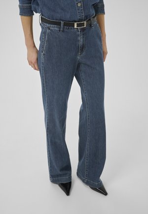 Persona tērpus tumši zilas džinsa bikses ar melnu jostu un melnas smailas kurpes, stāvot uz vienkārša pelēka fona.