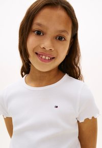 Jeune fille souriante aux longs cheveux bruns, portant un t-shirt blanc à manches courtes côtelé avec un petit logo sur la poitrine, devant un fond uni.