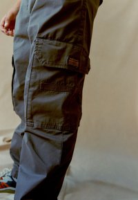 Pantaloni cargo grigio scuro con numerose tasche e un'etichetta patch sulla coscia. Realizzati in tessuto resistente, presentano cuciture tonali e una vestibilità rilassata.