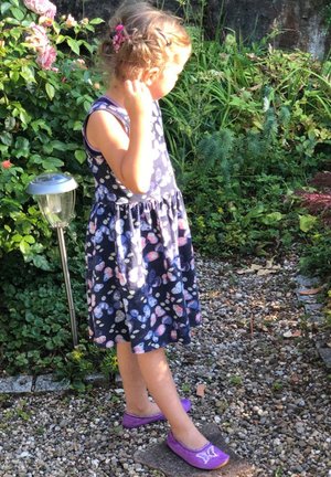 Navy dress with colorful butterfly print, flared skirt, and purple flat shoes with butterfly detail, standing on a gravel path.