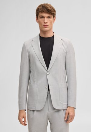 Giovane uomo con giacca da completo grigio chiaro sopra una camicia nera, in piedi davanti a uno sfondo grigio chiaro uniforme.