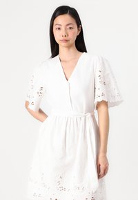 Vit klänning med V-ringning, korta ärmar med blommiga utskärningar, ett knytbälte i midjan och en texturerad fåll med broderade detaljer.