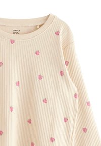 Kermanvärinen ribbattu pitkähihainen paita, jossa on pieniä vaaleanpunaisia mansikkakuvioita, pyöreä pääntie ja pehmeä tekstuuri.