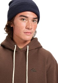 Sudadera con capucha marrón y cordones blancos, llevada sobre una camiseta verde. Gorro de punto azul en la cabeza. Piel con pecas.