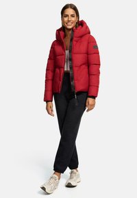 Red puffer jacket with a hood, featuring a zipped front and black accents. Paired with black joggers and light-colored sneakers.