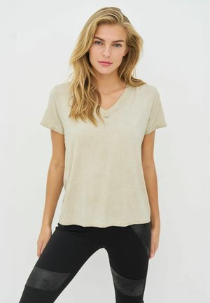 Lyshåret kvinde med løst hår iført en beige V-hals kortærmet skjorte og sorte leggings med grå lapdetaljer, stående mod en hvid baggrund.