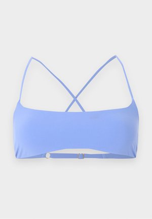 Grobe Frau mit dunkler Haut, die einen hellblauen Bikini trägt, barfuß steht und eine Hand auf der Hüfte vor weißem schlichten Hintergrund hat.