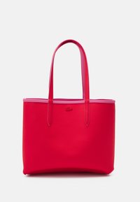 Sac fourre-tout rouge en matériau synthétique texturé, avec de longues poignées roses et un logo de marque discret embossé à l'avant.