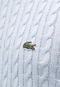 Tissu bleu clair en tricot torsadé avec un petit logo crocodile vert brodé, présentant une bouche ouverte avec une langue rouge.