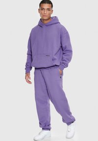 Lilla hættebluse og matchende joggers, lavet af blødt stof. Hætteblusen har en frontlomme; joggersne har lynlåslommer på siden.