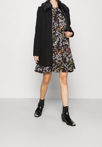 Femme portant un manteau noir avec un col en fourrure sur une robe à fleurs et des bottines noires avec embouts en métal, se tenant contre un fond uni.
