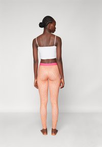 Ferskenfargede leggings med et bølgete oransje mønster og rosa midje, sammen med en hvit crop-top. Modellen står barføtt og vender bort.