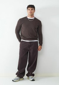 Brauner Pullover, der über einem weißen Hemd getragen wird, kombiniert mit dunkelgrauen, lockeren Hosen und silbernen Sneakers. Lässiges Design mit strukturierten Materialien.