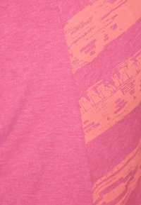 Różowa koszulka wykonana z lekkiego materiału, z diagonalnym pomarańczowym wzorem graficznym oraz teksturowanymi liniami po jednej stronie.