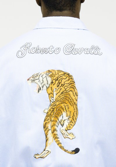 Dos d'une chemise bleu clair avec "Roberto Cavalli" brodé au-dessus d'un tigre rugissant détaillé, brodé en fil jaune, noir et blanc.