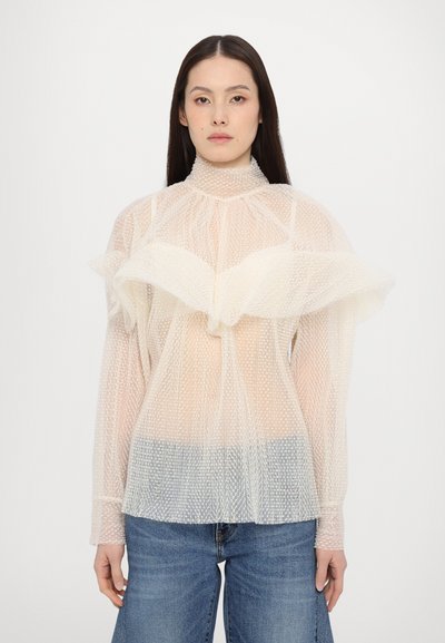 Haut transparent de couleur crème avec un col haut, motif de pois texturé et design à volants superposés. Manches longues avec des poignets élastiques.