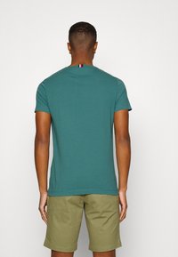 Mand med kort hår iført en teal t-shirt med et rød, hvidt og blåt mærke på ryggen af halsudskæringen og olivengrønne shorts, der står op ad en hvid væg.