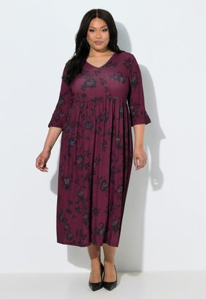 Donna in piedi che indossa un vestito lungo a fiori viola scuro con maniche a tre quarti e scarpe nere con tacchi alti su uno sfondo semplice.