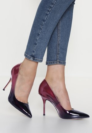 Mujer con vaqueros pitillo azules y zapatos de tacón alto brillantes en degrade púrpura con punta puntiaguda, de pie sobre una superficie blanca.