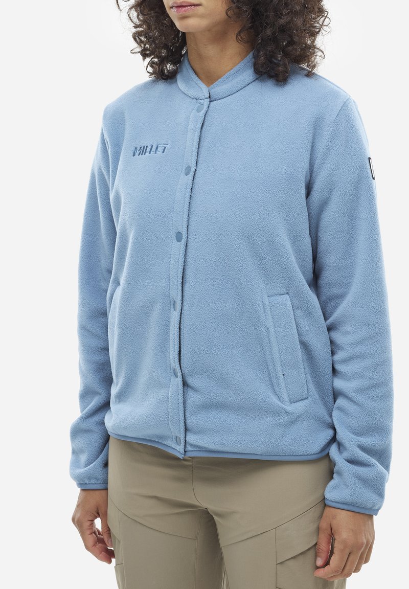 Veste en polaire bleu clair avec une fermeture à boutons, des poignets élastiques, des poches latérales et un logo sur la poitrine. Texture douce et design simple.