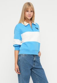 Joven mujer con cabello rubio vestida con una sudadera de cremallera a rayas azul y blanca y jeans azules, de pie con una mano en el bolsillo.