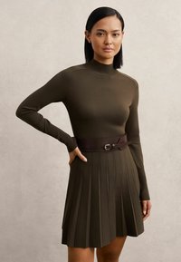 Olívazöld kötött ruha hosszú ujjakkal, magas gallérral és plisszírozott szoknyával. Jellemzője egy sötétbarna bőr öv, amely csattal kiegészített.