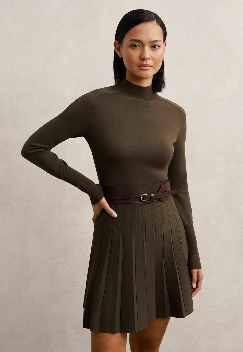 Olívazöld kötött ruha hosszú ujjakkal, magas gallérral és plisszírozott szoknyával. Jellemzője egy sötétbarna bőr öv, amely csattal kiegészített.