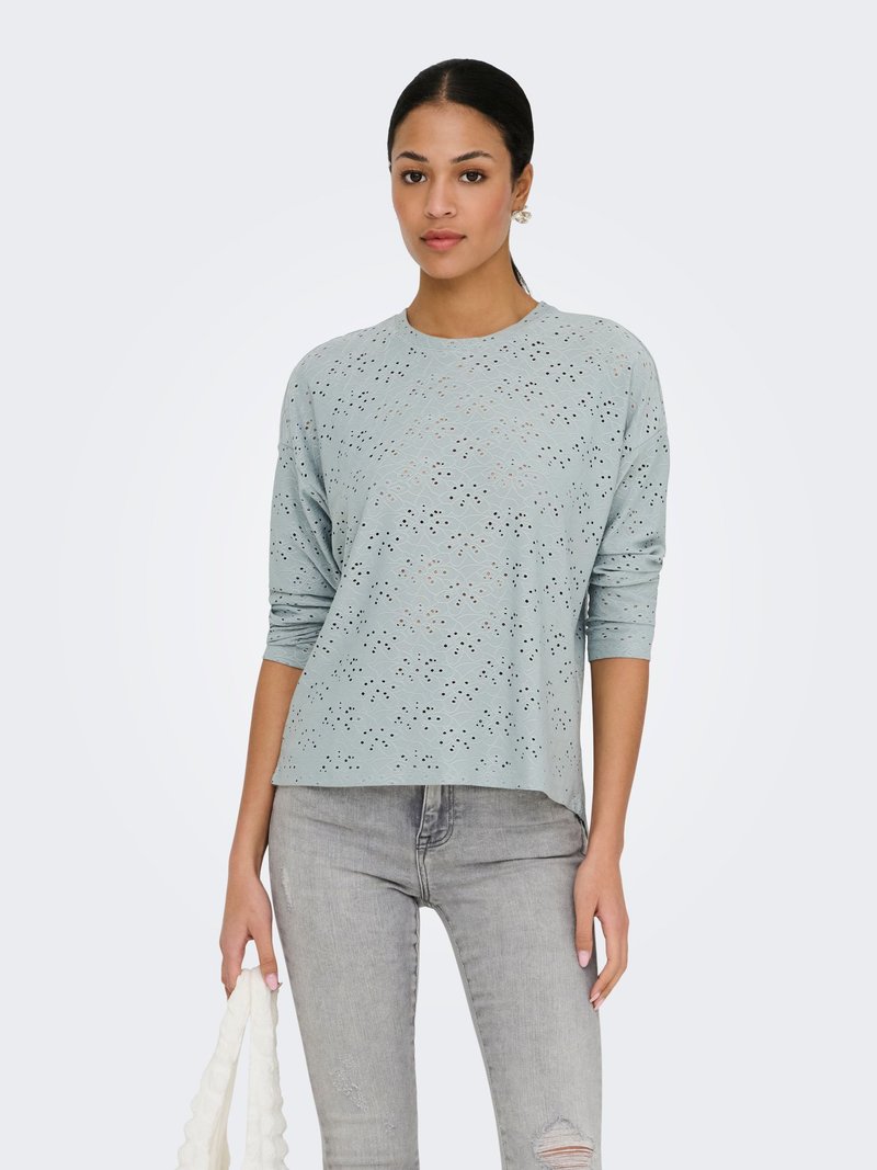 Γυναίκα που φοράει ένα ανοιχτό γαλάζιο μακρυμάνικο τοπ με σχέδιο από τρυπητό ύφασμα και ανοιχτό γκρι σκισμένο στενό τζιν, κρατώντας μια λευκή τσάντα.