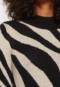 Svart och beige randig stickad tröja med en texturerad finish. Har en svart ribbad rund hals och kontrasterande djärva zebra-liknande mönster.