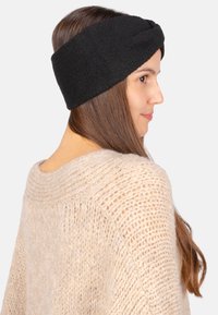 Bandeau en polaire noire avec un design à l'avant torsadé. Texture lisse, style ajusté, porté sur de longs cheveux bruns, associé à un pull en tricot beige.