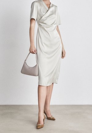 Frau trägt ein weißes Wickel-Midikleid, hält eine beige Handtasche und trägt braune High Heels mit goldener Kettendetails.