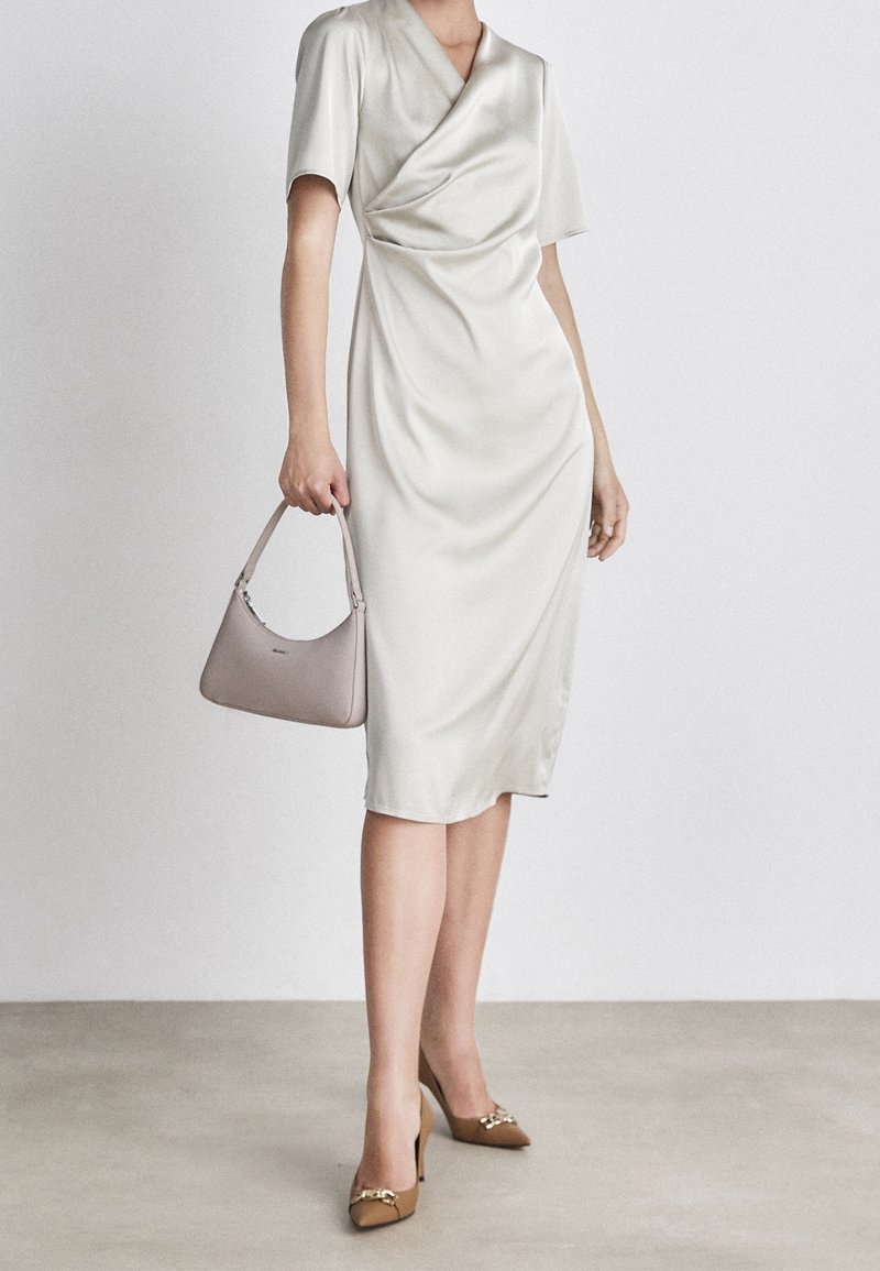Femme portant une robe midi portefeuille blanche, tenant un sac à main beige et portant des talons hauts marron avec un détail chaîne dorée.