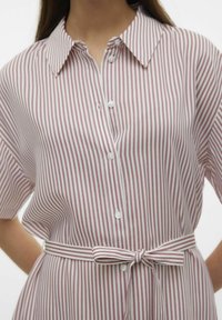 Camisa a rayas con cuello, cierre de botones y mangas cortas. Los colores son rosa claro y blanco, con un cinturón de lazo a juego en la cintura.
