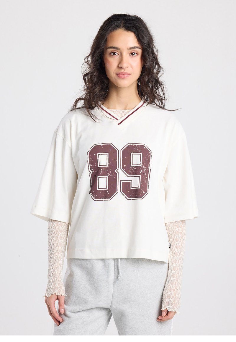 Witte cropped t-shirt met rode bies en groot distressed bordeauxkleurig nummer 89. Model draagt een kanten longsleeve eronder en grijze joggingbroek.