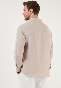 Hellbeige Langarmhemd mit geradem Kragen, strukturiertem Stoff und Ein-Knopf-Manschetten, kombiniert mit weißen Hosen.
