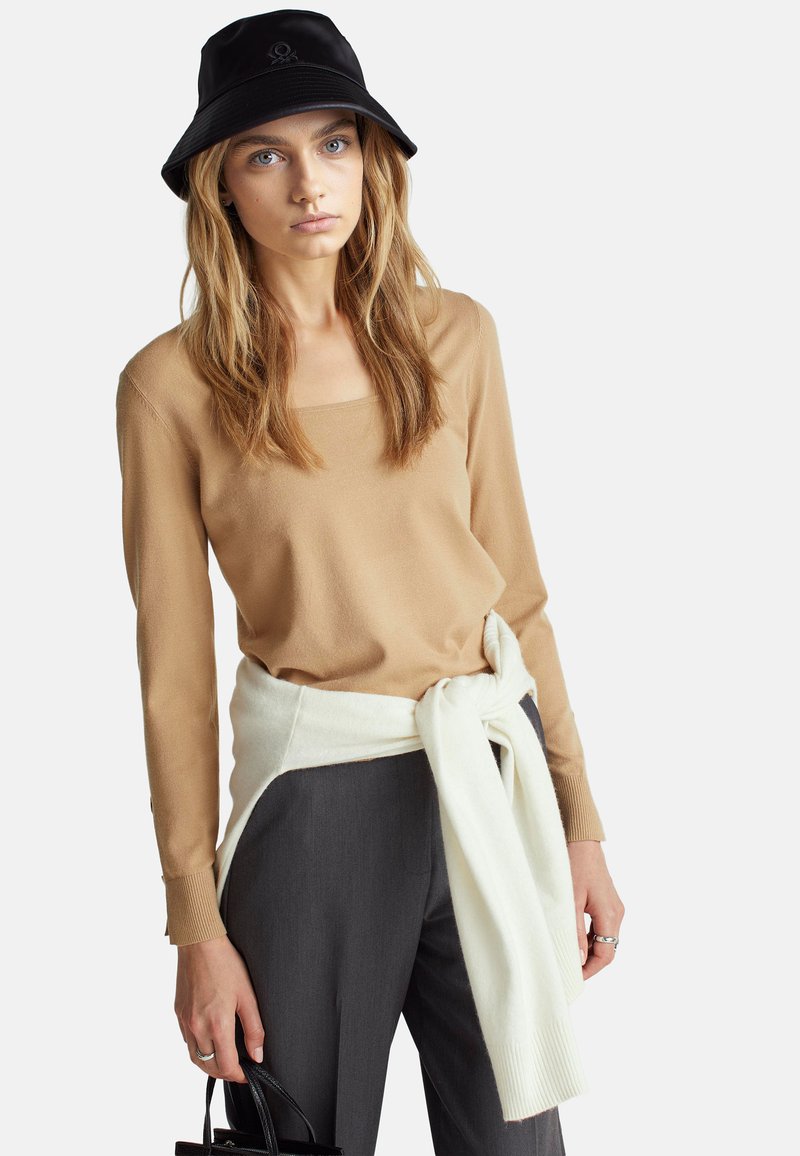 Beżowa bluzka z długim rękawem i kwadratowym dekoltem, biała sweter związany w talii, szare dopasowane spodnie oraz czarna kapelusz typu bucket z logo.