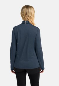 Frau mit blonden Haaren, die ein langärmliges navy-blaues Oberteil und schwarze Hosen trägt, steht mit dem Rücken zu einem schlichten weißen Hintergrund.