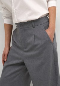 Pantalon gris sur mesure avec une ceinture structurée, des plis à l'avant et des poches latérales, fabriqué dans un tissu lisse. Coupe classique à jambes droites.