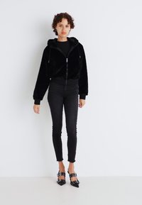 Zwarte nep-bont zip-up hoodie met voorzakken, gecombineerd met zwarte skinny jeans en zwarte gestifte hakken. Soepele textuur en relaxte pasvorm.