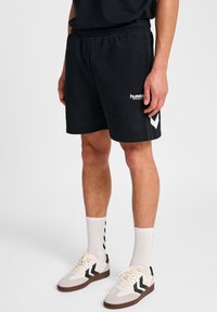 Man in zwarte Hummel-shorts, witte hoge sokken met zwarte chevrons, en witte sneakers met zwarte chevrons op een effen achtergrond.