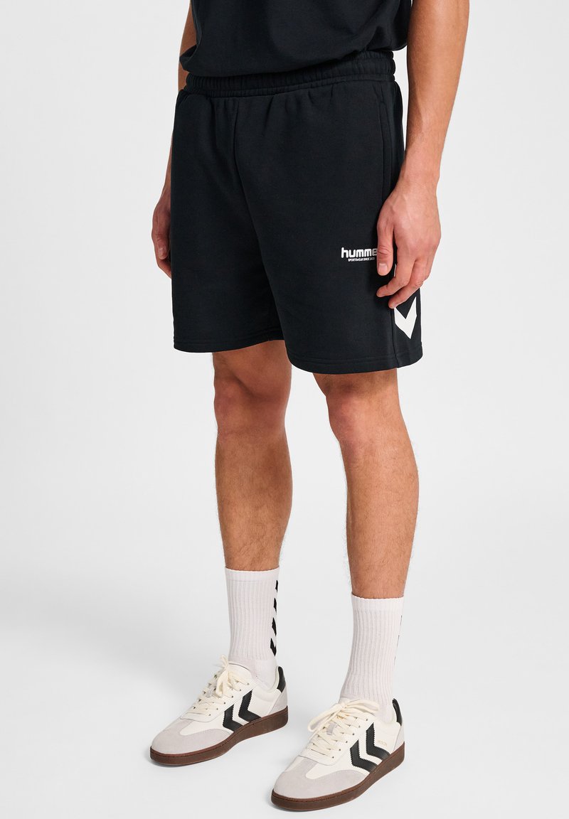 Man in zwarte Hummel-shorts, witte hoge sokken met zwarte chevrons, en witte sneakers met zwarte chevrons op een effen achtergrond.