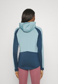 Huvtröja i ljus och mörk blå med långa ärmar, med en rundad nederkant och kontrastfärgad blockdesign. Slät textur.