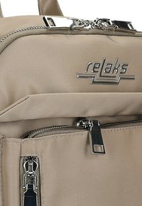 Beżowy plecak z tkaniny z metalowymi elementami w kolorze srebrnym, wyposażony w wiele zapinanych na zamek przegród oraz wyraźnym logo z przodu. Gładka faktura.