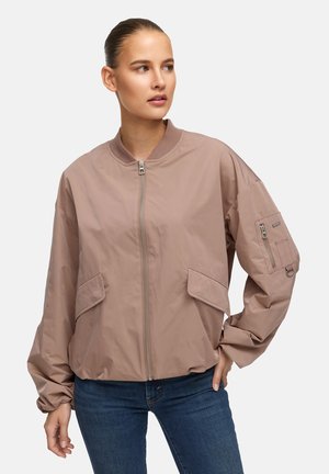 Frau trägt eine hellbraune Bomberjacke mit Reißverschluss, Klapptaschen und einer Reißverschlusstasche am Ärmel, kombiniert mit blauen Jeans, blickt zur Seite.